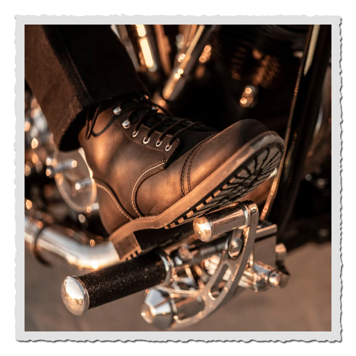 Nahaufnahme eines schwarzen Stiefels auf Motorrad-Fußraste, warmes Licht, chromige Teile im Hintergrund.